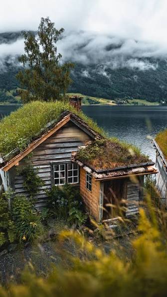 Gammal tømmerhytte med torvtak ved vatn og fjell